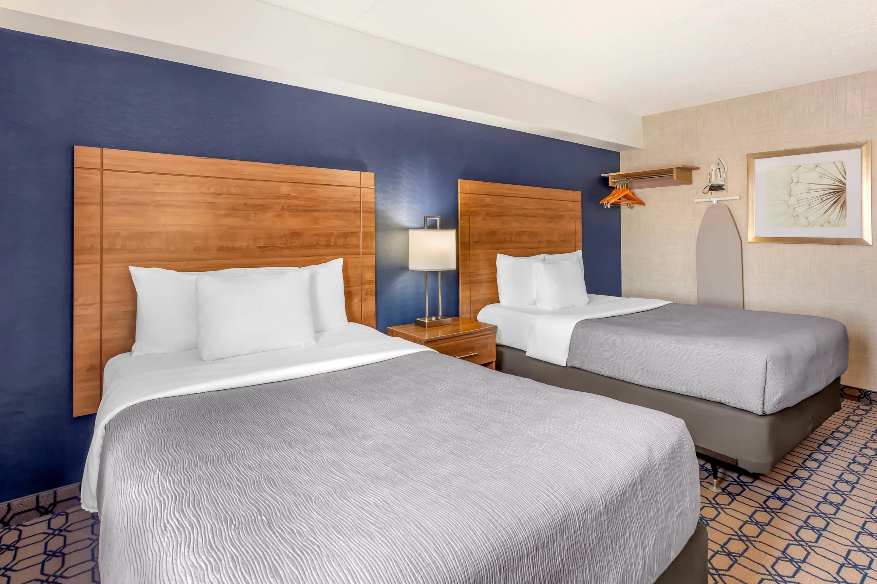 A hotel room with two beds, a nightstand, a lamp, a coat rack, and a framed picture on the wall.