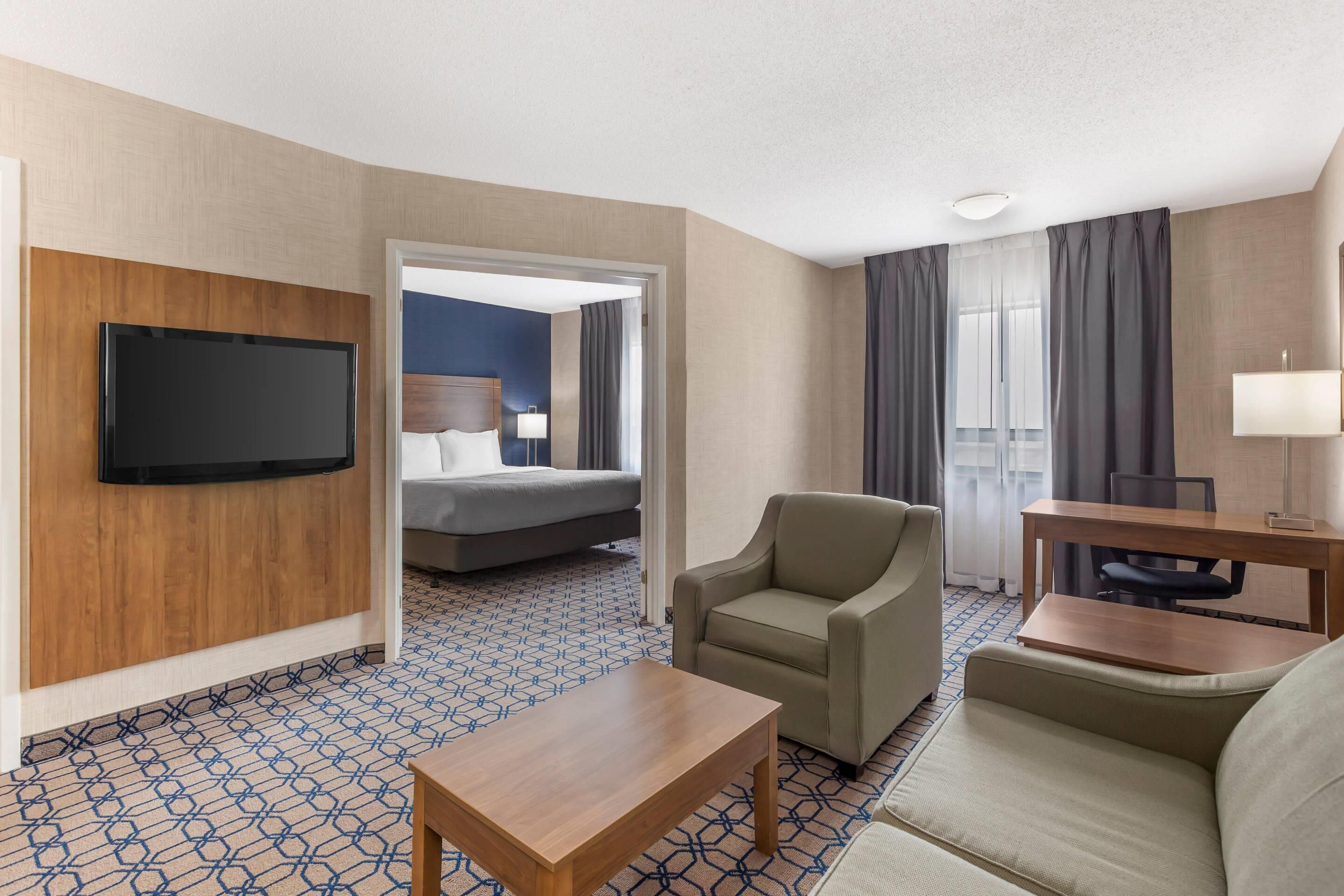 Quarto de hotel com televisão de tela plana na parede, um sofá, uma mesa pequena, uma cama com cabeceira e uma janela com cortinas.