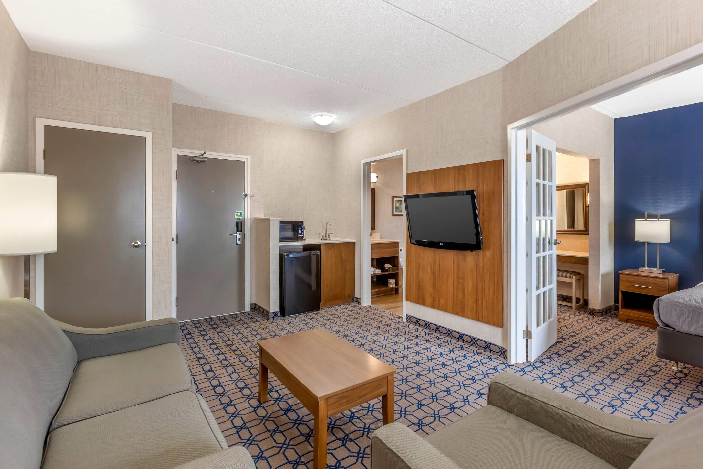 A hotel room with a sofa, a small table, a TV mounted on the wall, a desk with a chair, and a kitchenette area.