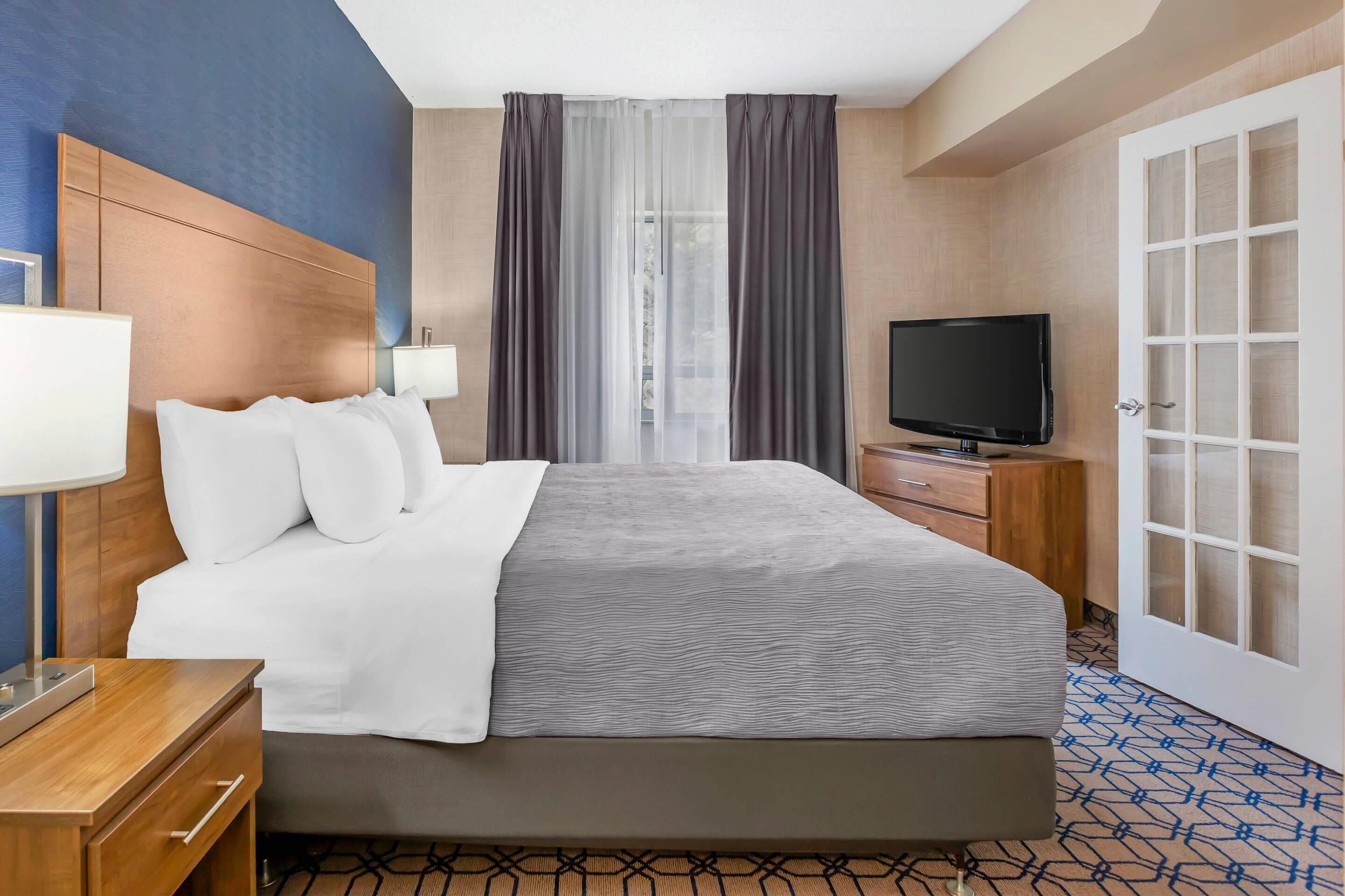 Habitación de hotel con cama, mesita de noche, lámpara, televisor y una puerta con panel de vidrio.