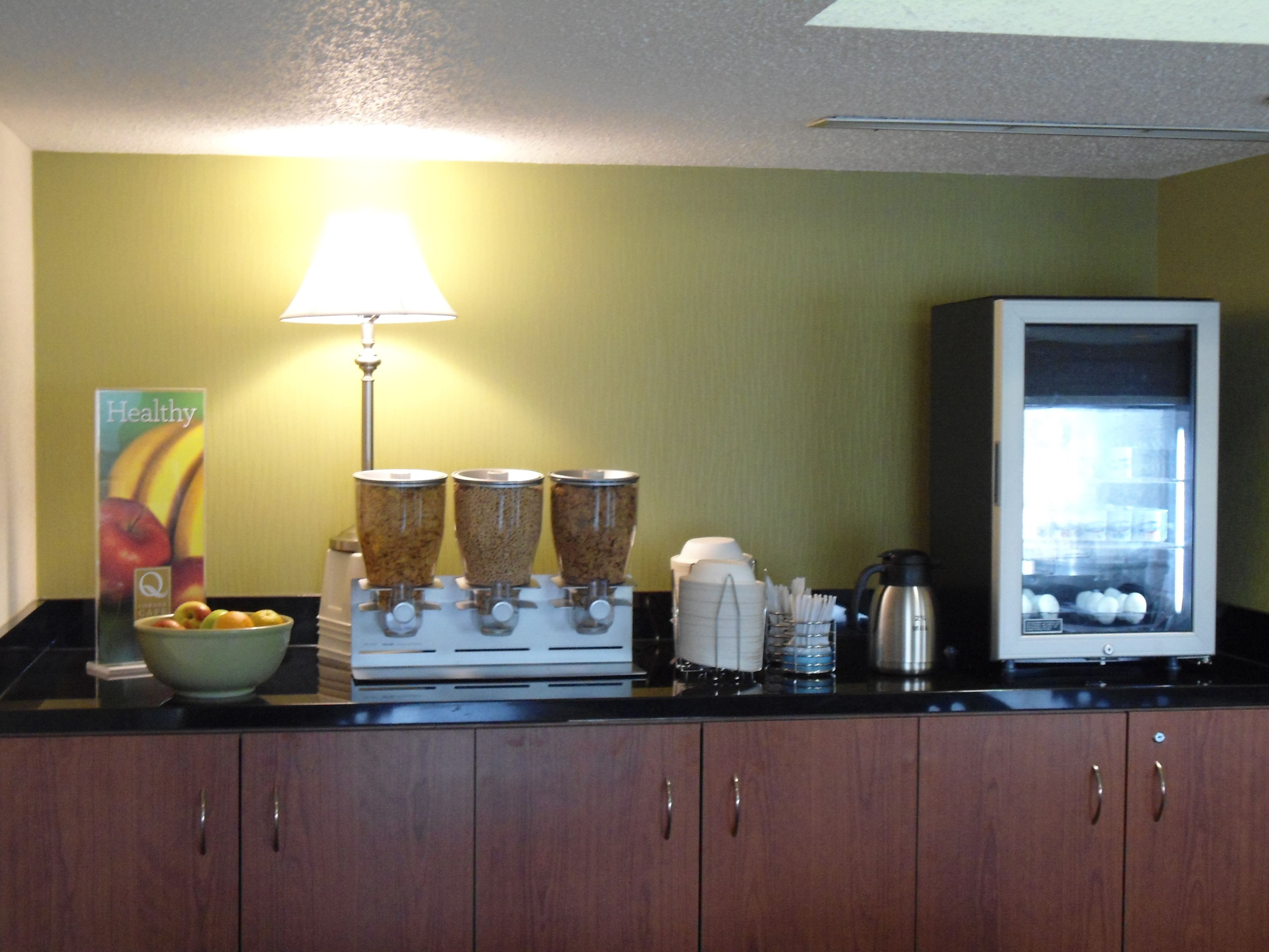 A kitchen with a coffee maker, a bowl of fruit, a coffee dispenser, and a refrigerator.