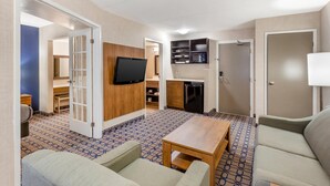 A hotel room with a sofa, a coffee table, a TV mounted on the wall, a kitchenette with a microwave, and a door leading to a bathroom. - Quality Suites Whitby (Whitby)