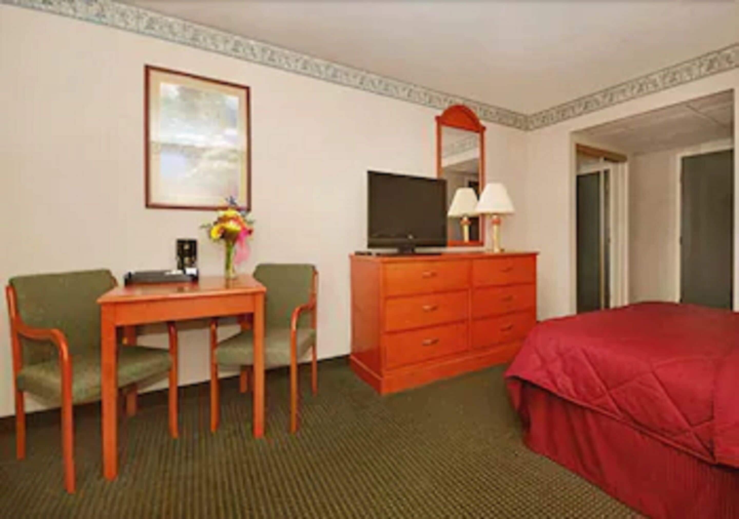 A hotel room with a bed, a desk with a chair, a television, a dresser, and a framed picture on the wall.