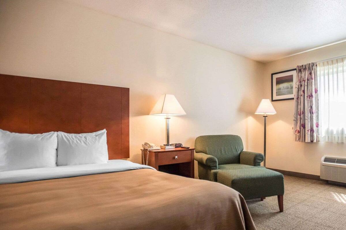 Ein Hotelzimmer mit einem großen Bett, einem Sessel, einem Nachttisch mit Lampe und einem Fenster mit Vorhängen.