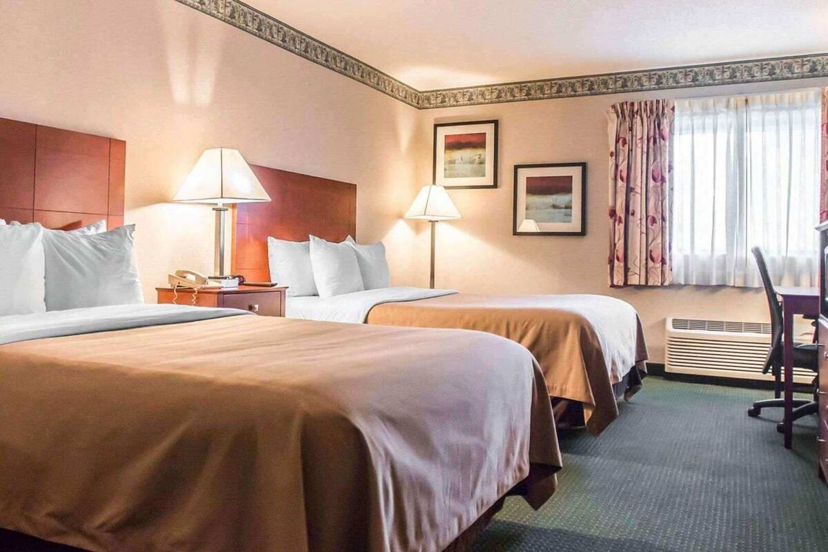 Ein Hotelzimmer mit zwei Betten, einem Schreibtisch, einem Stuhl, einem Telefon und einem Fenster mit Vorhängen.