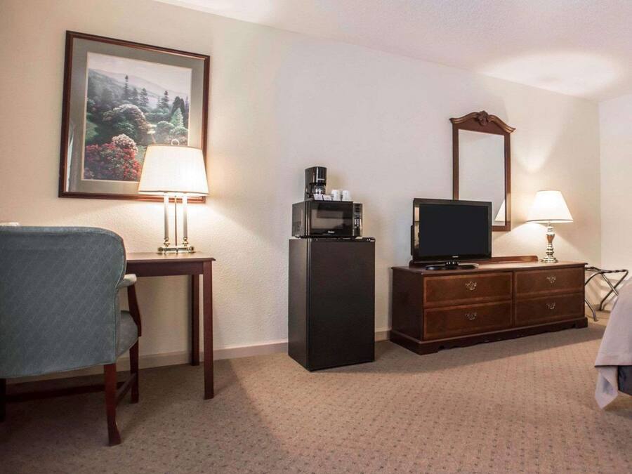 A hotel room with a bed, a desk with a lamp, a refrigerator, a television, and a dresser.