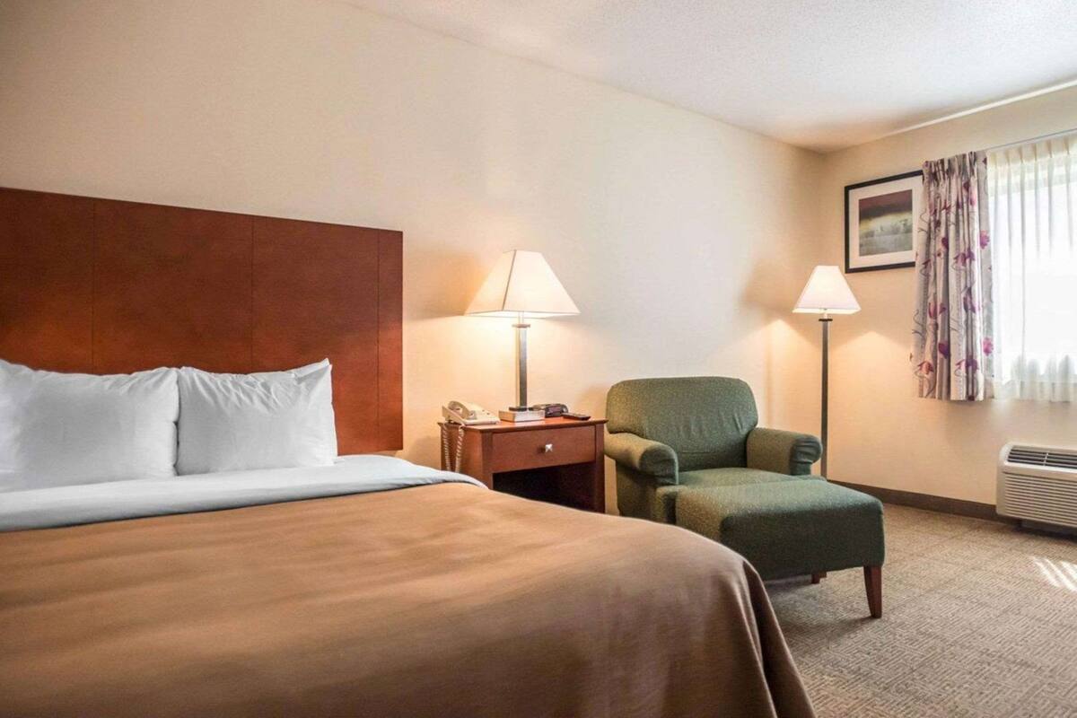 Ein Hotelzimmer mit Bett, Stuhl, Nachttisch, Lampe, einem Bild an der Wand und einem Fenster mit Vorhängen.