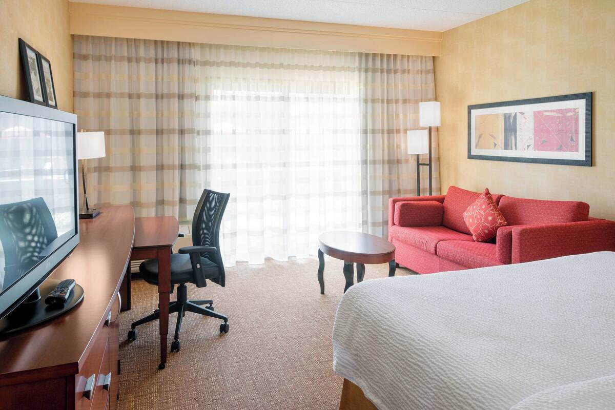 A hotel room with a bed, a desk with a computer, a chair, a red sofa, and a large window with curtains.