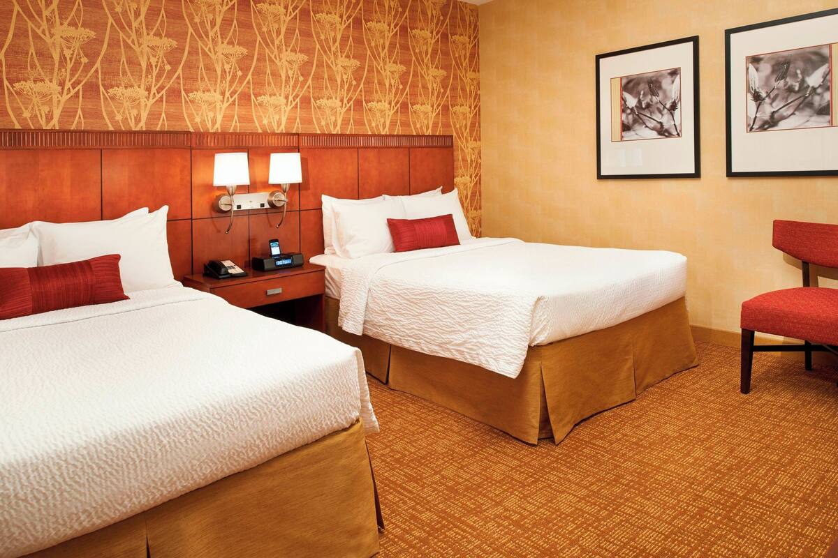 A hotel room with two beds, a wooden headboard, a nightstand with a phone and lamp, and a red chair.