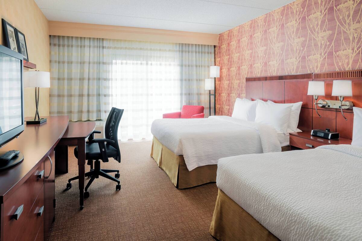 A hotel room with two beds, a desk, a chair, and a TV.