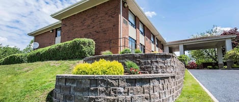 A two-story brick building with a flat roof, a covered entrance, and a landscaped area with a stone retaining wall and various shrubs.