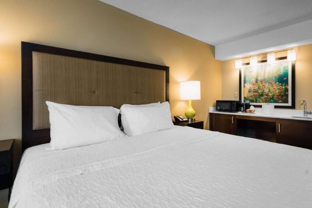 A hotel room with a large bed, a nightstand with a lamp, a microwave, and a painting on the wall.
