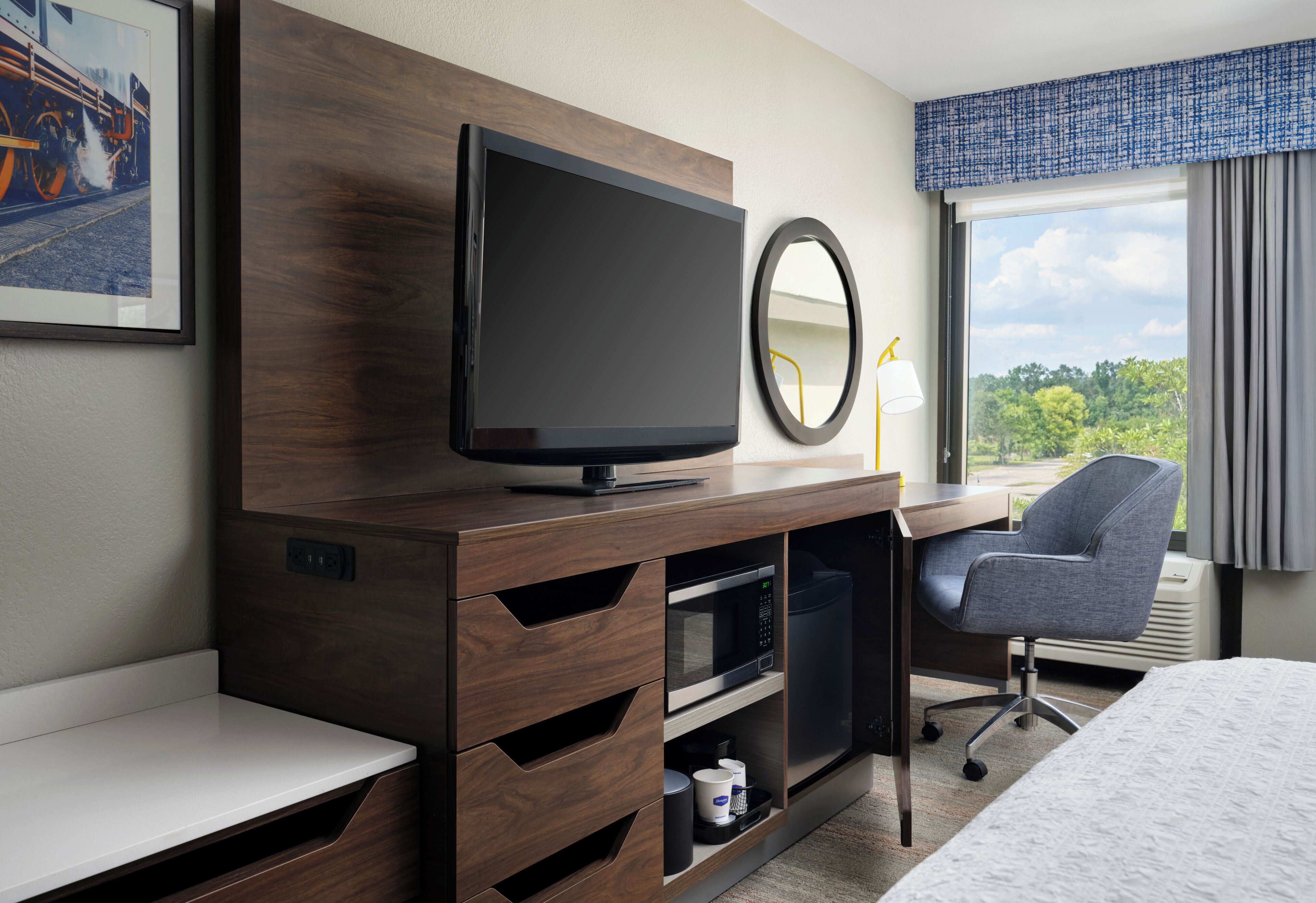 Une chambre d’hôtel équipée d’une télévision, d’un bureau, d’une chaise, d’un lit, d’une fenêtre avec des rideaux et d’un tableau encadré au mur.