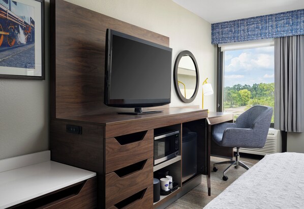 A hotel room with a TV, a desk, a chair, a bed, a window with curtains, and a framed picture on the wall.