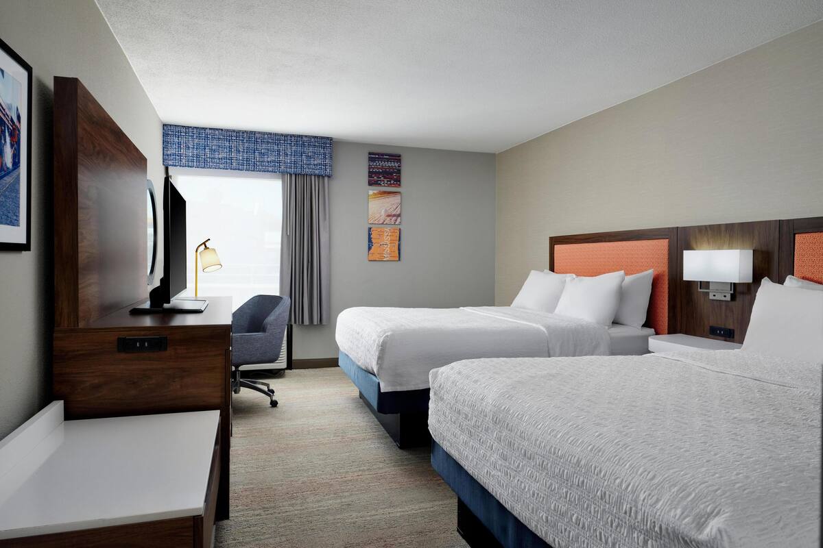 A hotel room with two beds, a desk, a chair, and a television.