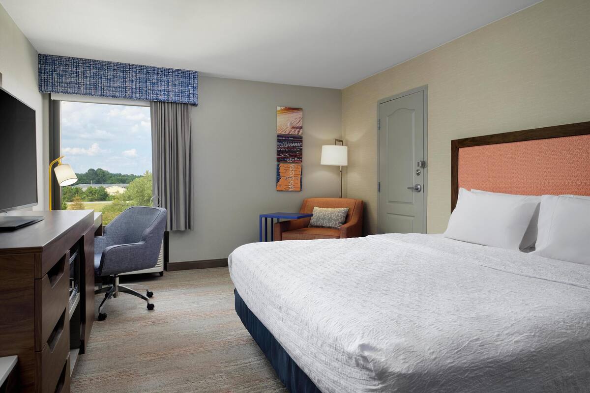 A hotel room with a large bed, a desk with a chair, a lamp, and a view of the outdoors.