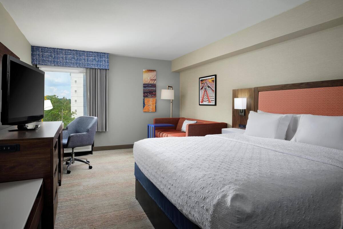 A hotel room with a bed, a desk with a TV, a chair, a sofa, and artwork on the walls.