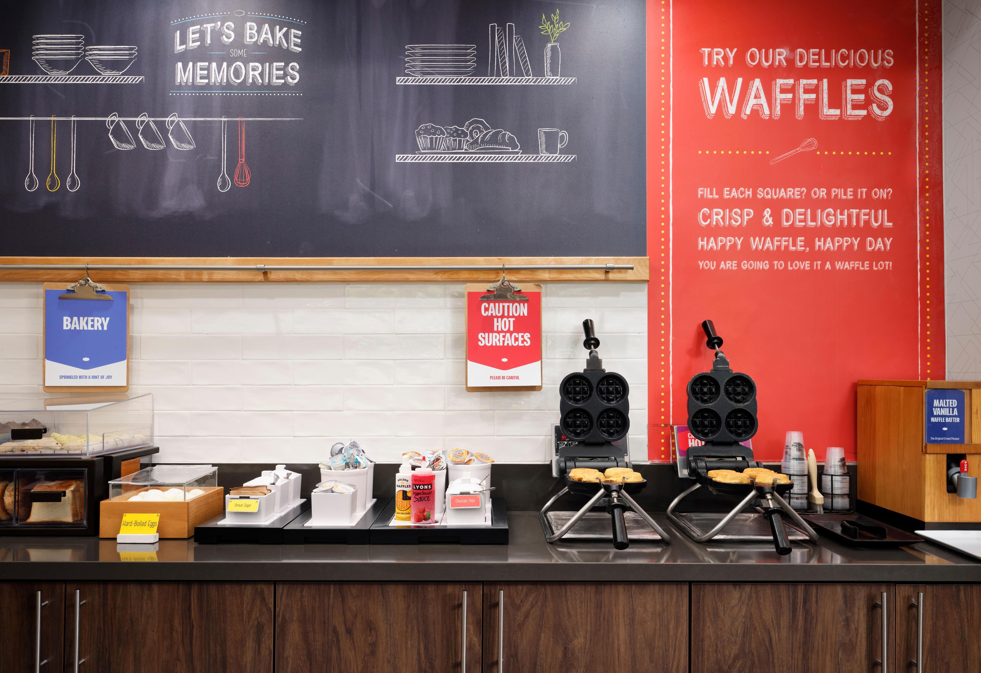 Balcão de pastelaria com equipamentos para fazer waffles, uma lousa com dicas de confeitaria e diversos produtos de panificação expostos.