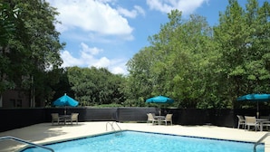 A swimming pool with a metal ladder, surrounded by a tiled deck. There are two blue umbrellas providing shade.