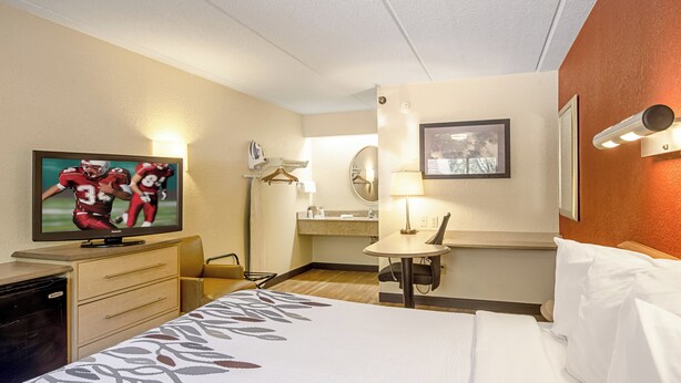A hotel room with a bed, a television displaying a sports game, a desk with a lamp, and a bathroom mirror. - Red Roof Inn St Clairsville - Wheeling West (St. Clairsville)