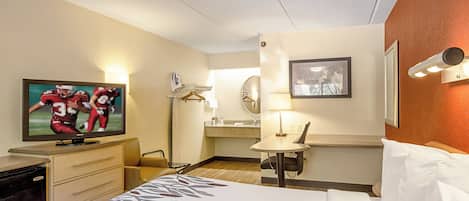 Habitación de hotel con cama, televisión mostrando un partido deportivo, escritorio con lámpara y espejo de baño.