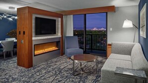 Une chambre d’hôtel moderne dotée d’une grande télévision, d’une cheminée, d’un balcon offrant une vue sur la ville, d’un canapé gris et d’une table basse ronde.