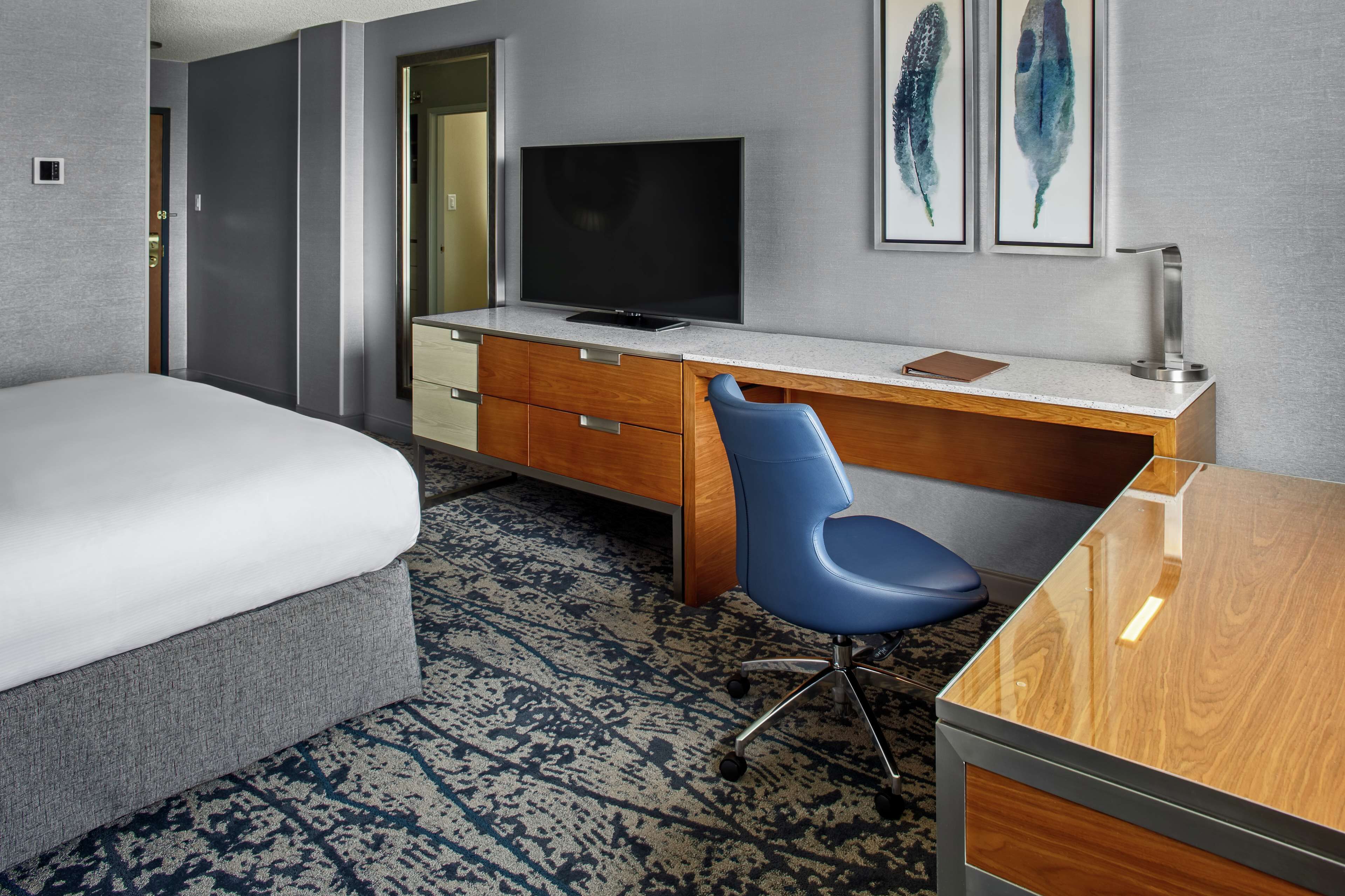 A hotel room with a bed, a desk with a chair, a television, and artwork on the wall.