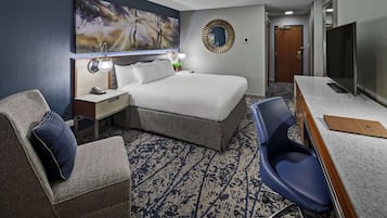 Ett hotellrum med en säng, ett skrivbord, en stol, en tv, en spegel och en tavla på väggen.