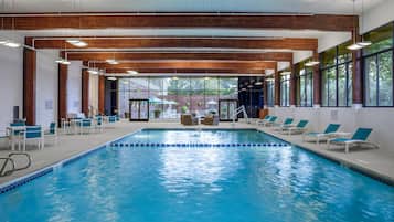 Una piscina interior con agua azul cristalina, rodeada de sillas y mesas de descanso, con ventanas amplias y vigas de madera en el techo.