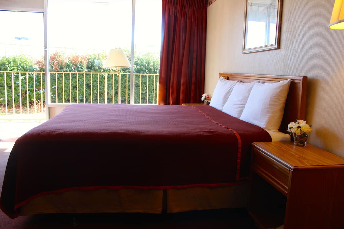 Habitación de hotel con una cama grande, una mesita de noche de madera, una mesita junto a la cama con una lámpara y una ventana con vistas a zonas verdes.