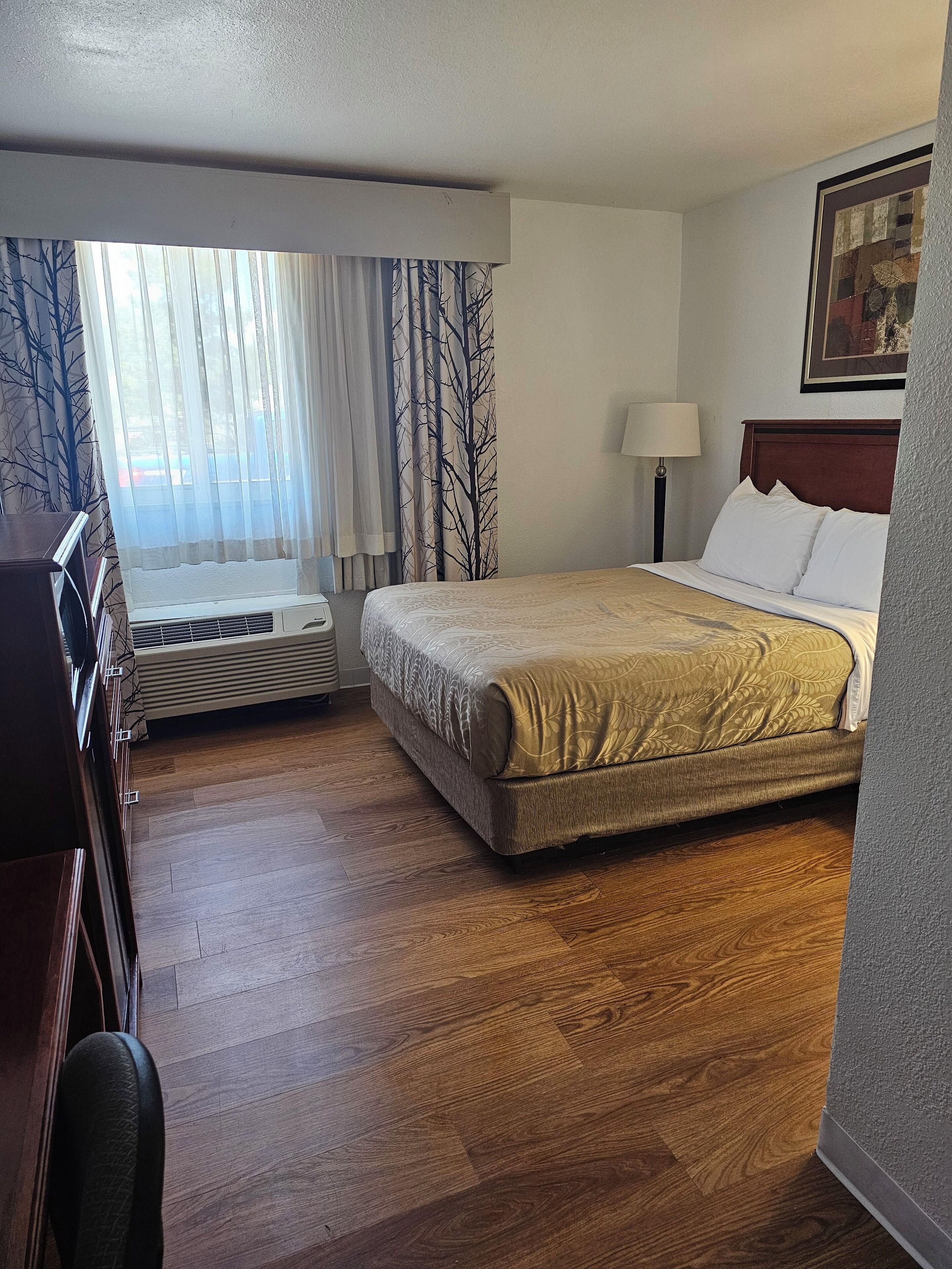 Habitació d'hotel amb un llit, escriptori, cadira, làmpada i finestra amb cortines.