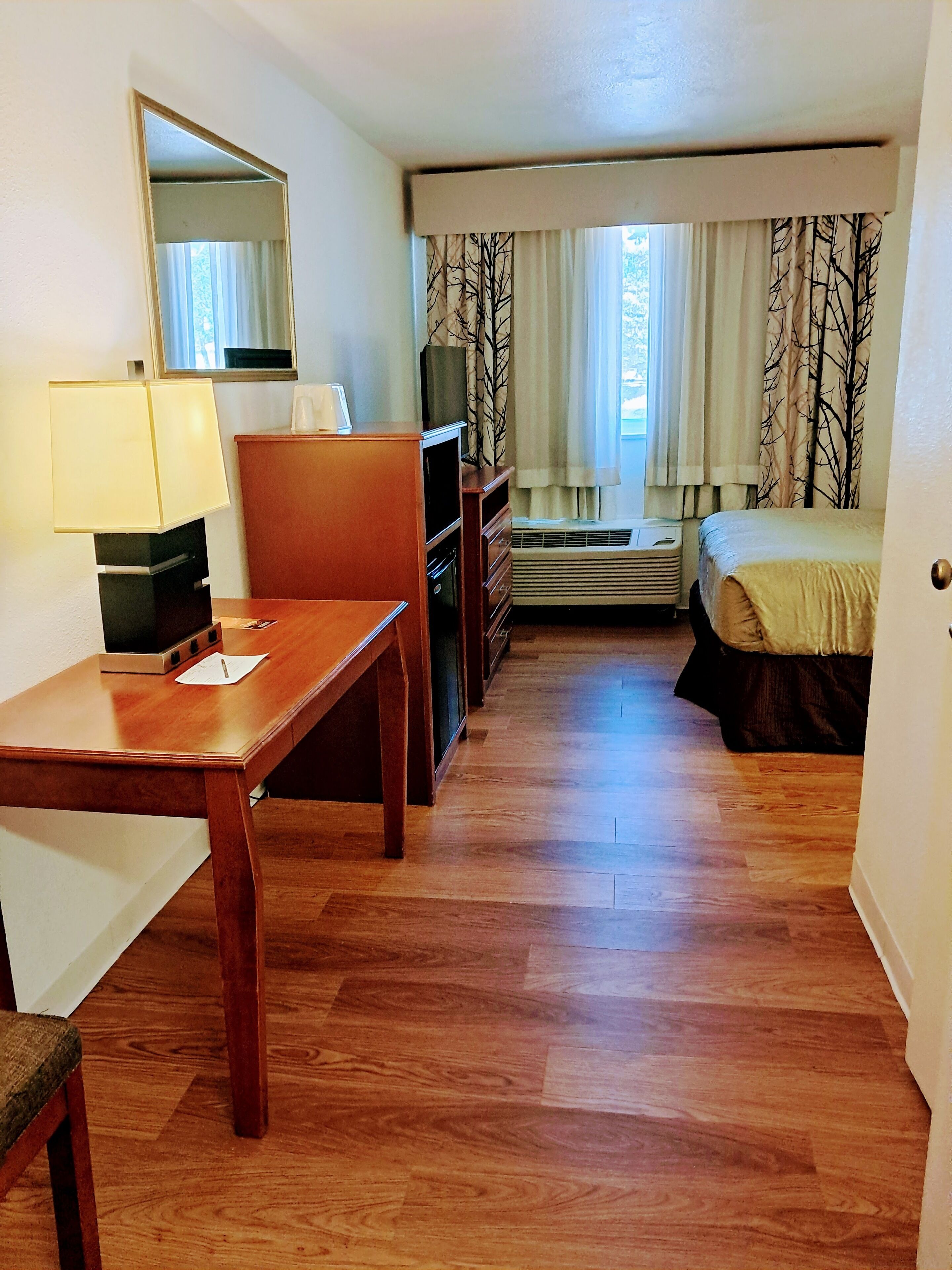 A hotel room with a wooden desk, a chair, a bed, a dresser, a mirror, and a window with curtains.