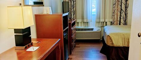 A hotel room with a wooden desk, a chair, a bed, a dresser, a mirror, and a window with curtains.