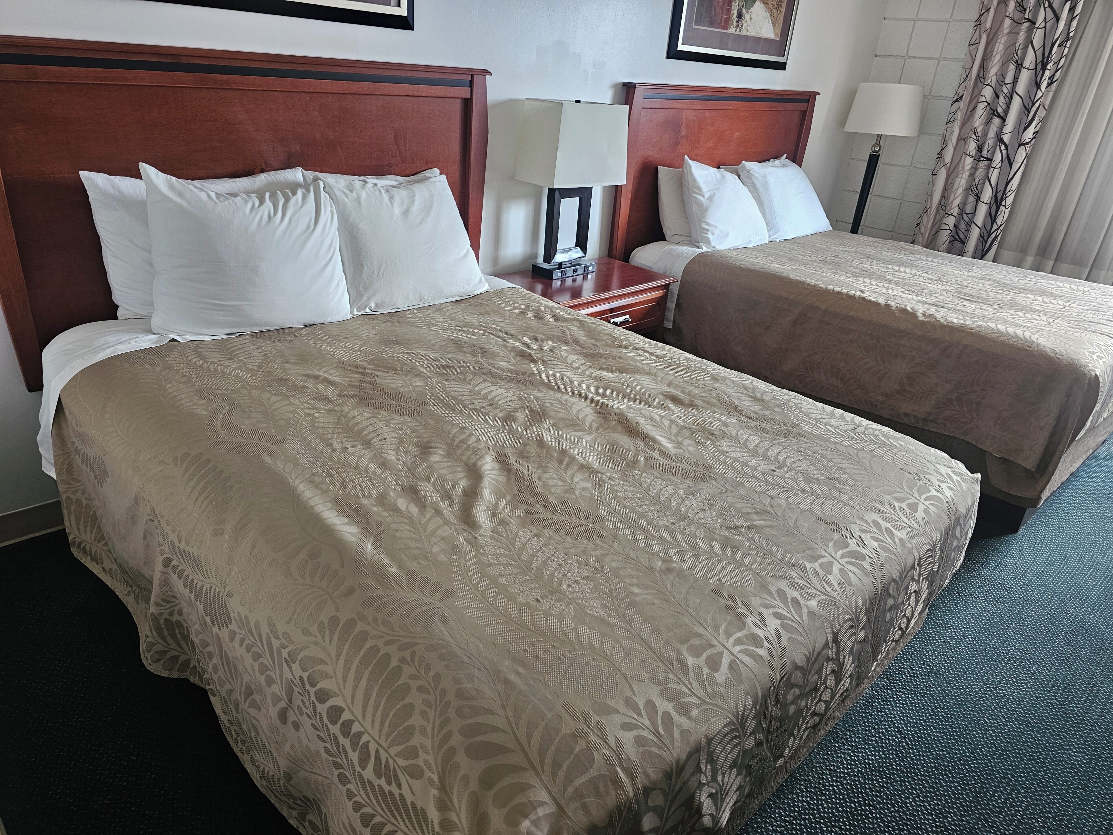 A hotel room with two beds, each with a wooden headboard and white bedding. There is a nightstand with a lamp and a small table between the beds.