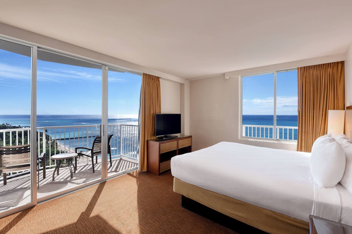 Une chambre d’hôtel avec un grand lit, un téléviseur posé sur un meuble en bois et un balcon offrant une vue sur l’océan.