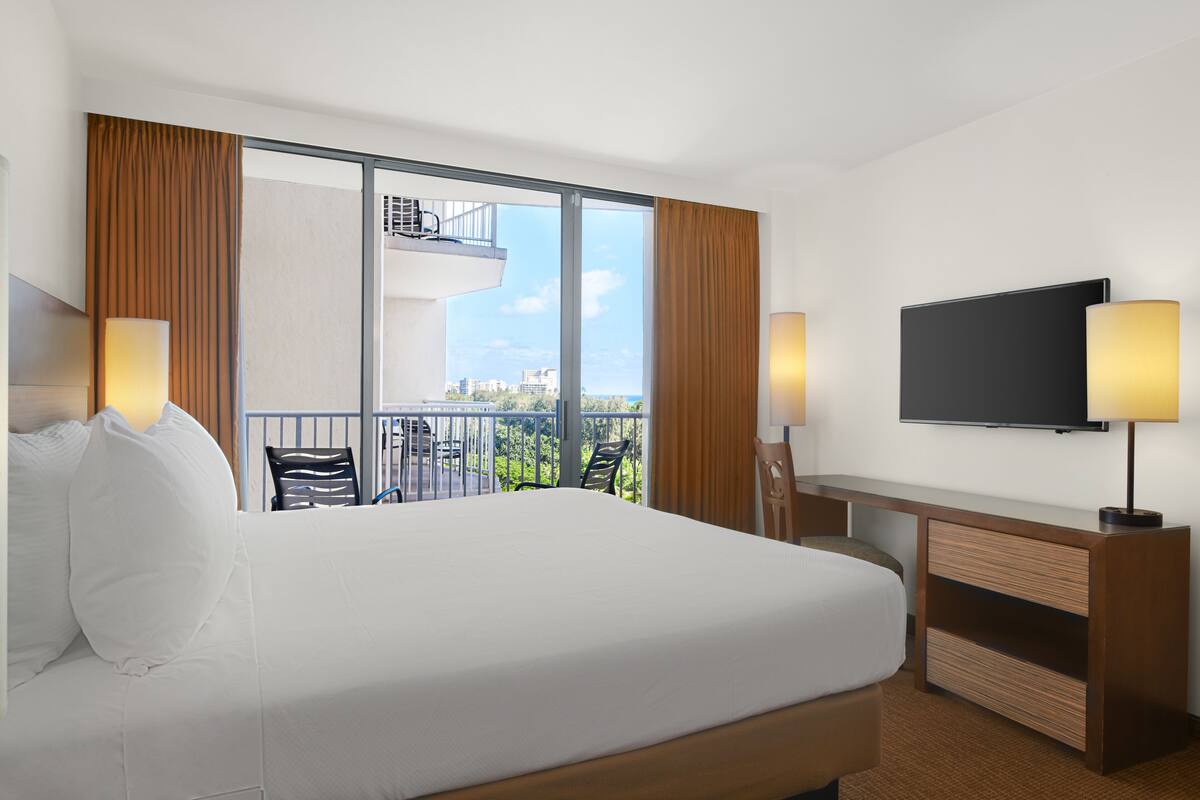 A hotel room with a large bed, a TV, a desk, and a balcony with a view of the city.