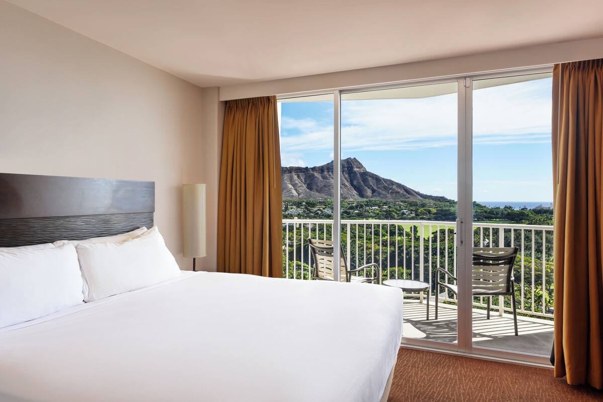 A hotel room with a large bed, a view of a mountain, and a balcony with chairs.