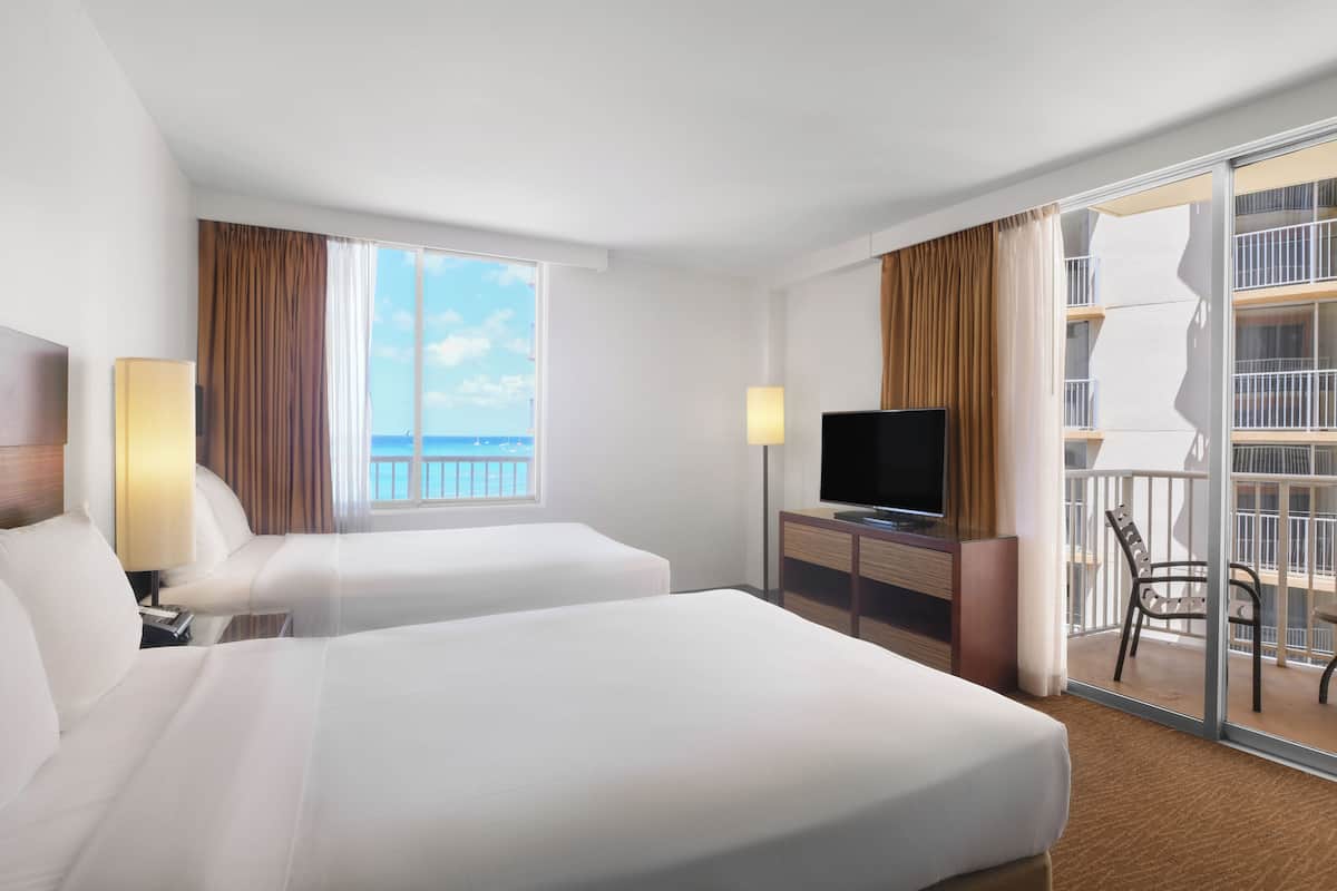 A hotel room with two beds, a TV, a desk, and a balcony with a view of the sea.