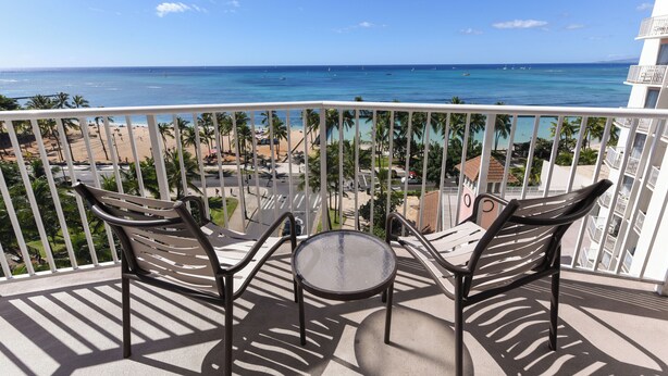 Un balcon avec deux chaises et une petite table, donnant sur une plage aux eaux d’un bleu limpide et bordée de palmiers.