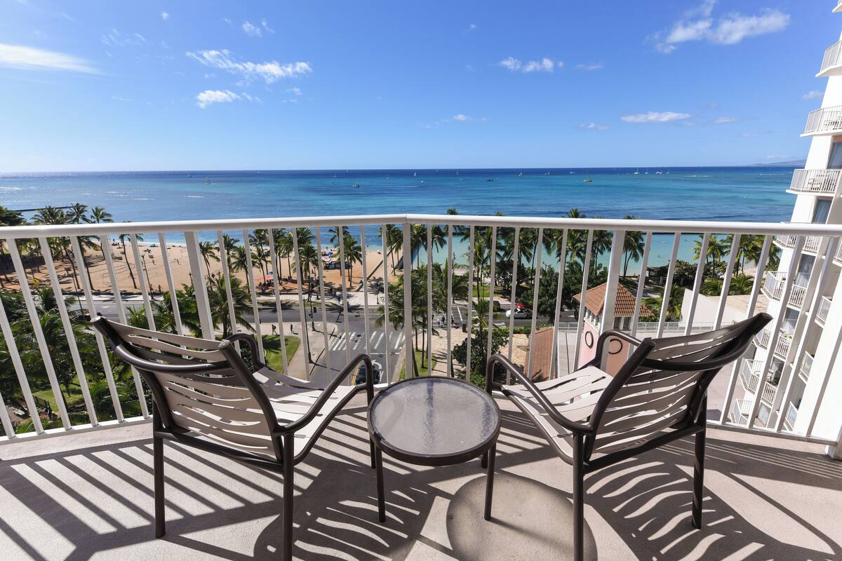 A balcony with two chairs and a small table, overlooking a beach with clear blue water and palm trees.
