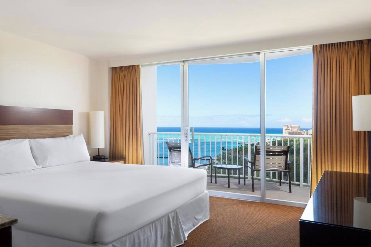 A hotel room with a bed, a desk, a chair, a table, and a balcony with a view of the ocean.