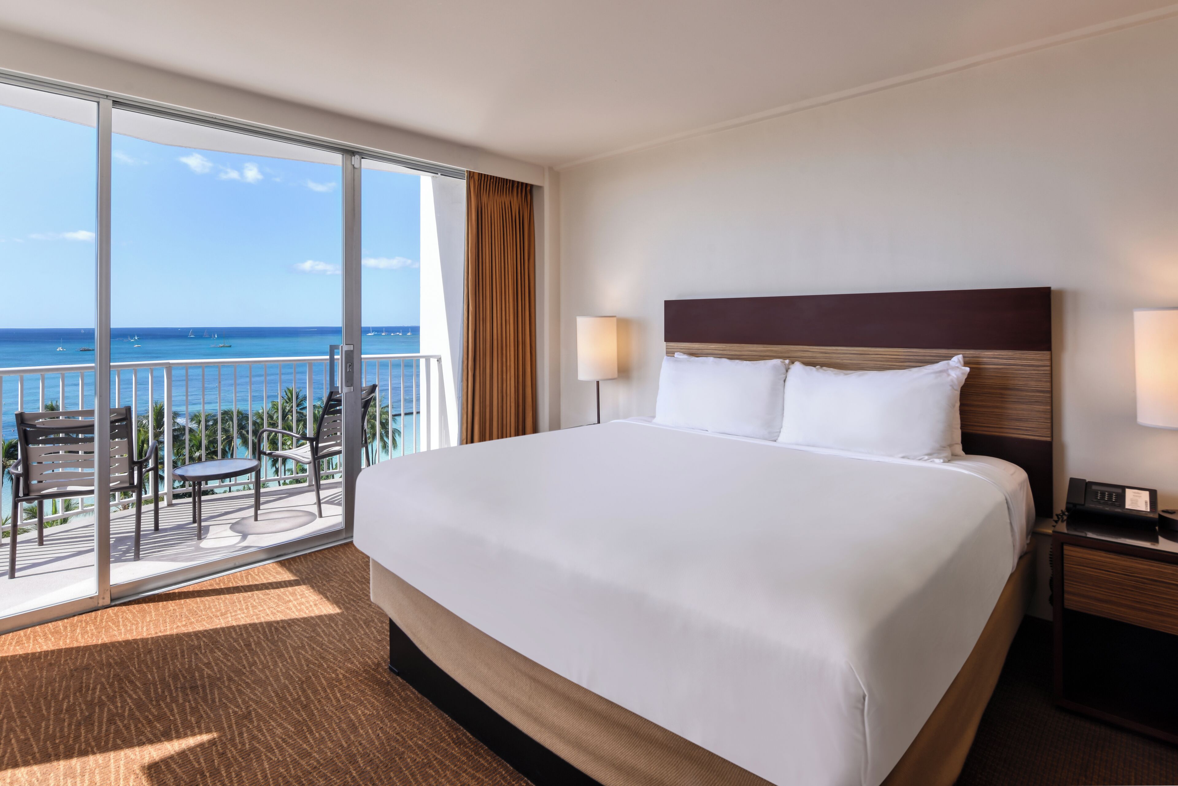 A hotel room with a large bed, a balcony with a view of the ocean, and a small table with chairs.