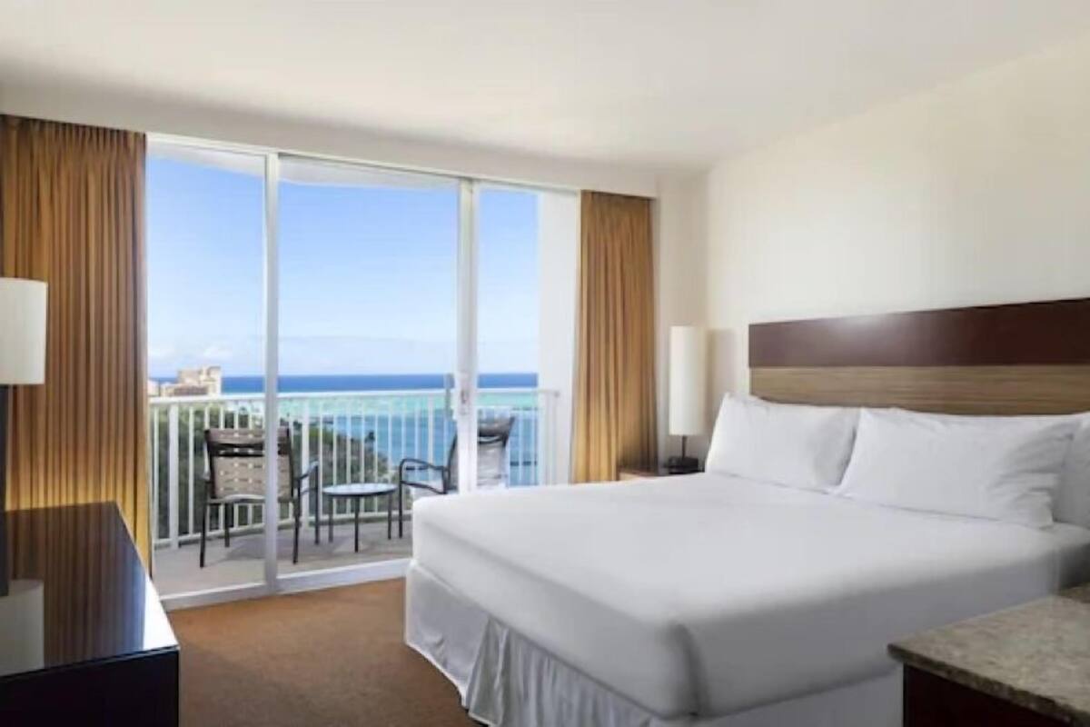 A hotel room with a large bed, a desk, and a balcony with a view of the ocean.