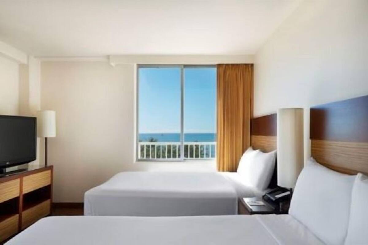 A hotel room with two beds, a TV, and a balcony with a view of the ocean.