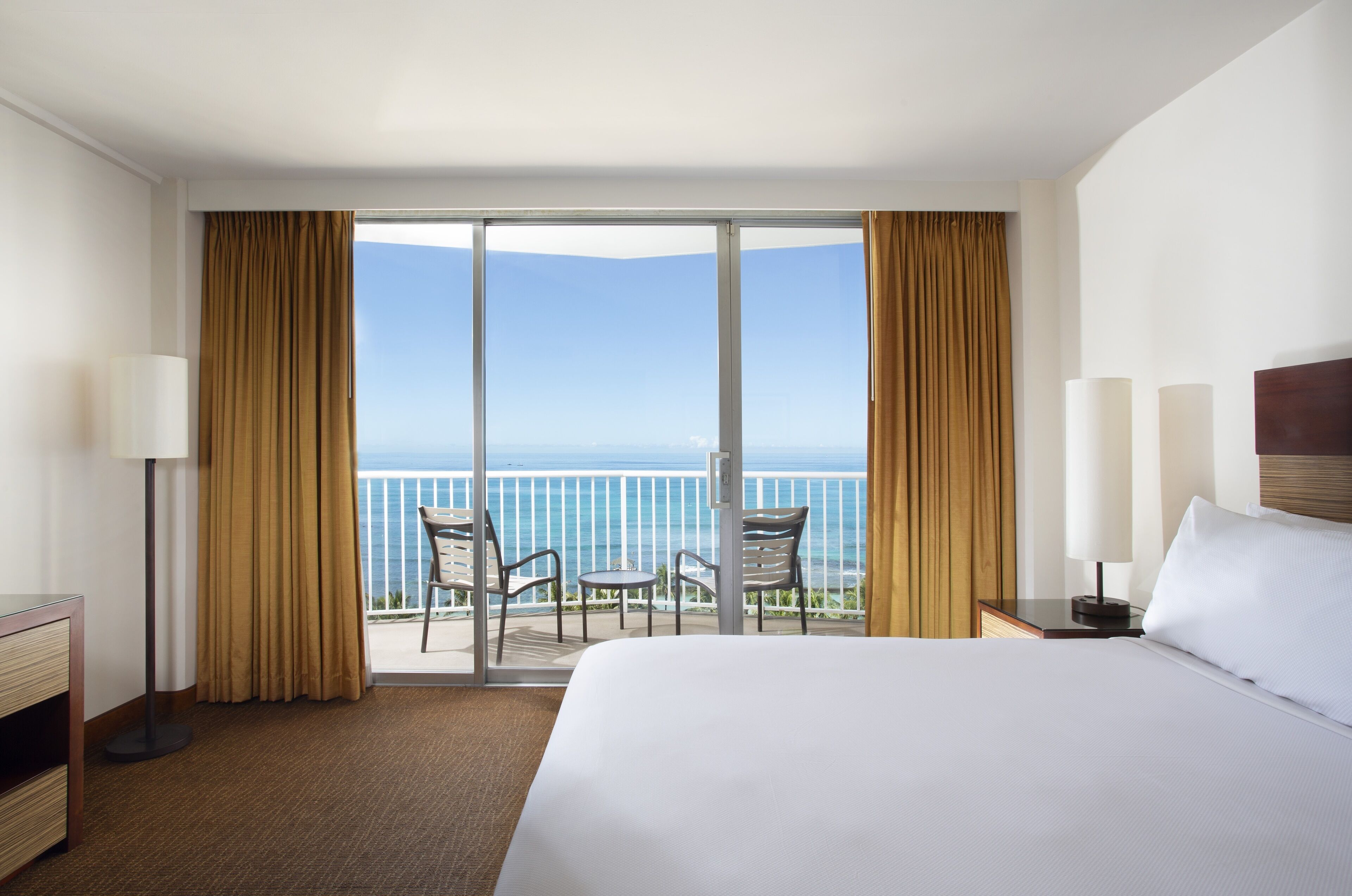 Una habitación de hotel con una cama grande, un escritorio, una silla, una mesita y vistas al océano a través de una puerta corrediza de vidrio.