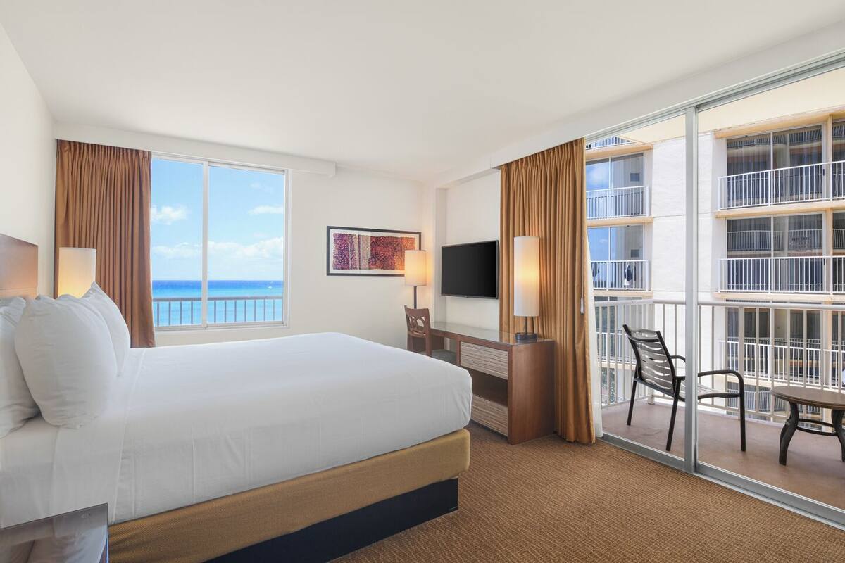 A hotel room with a bed, a desk, a chair, a TV, and a balcony with a view of the ocean.
