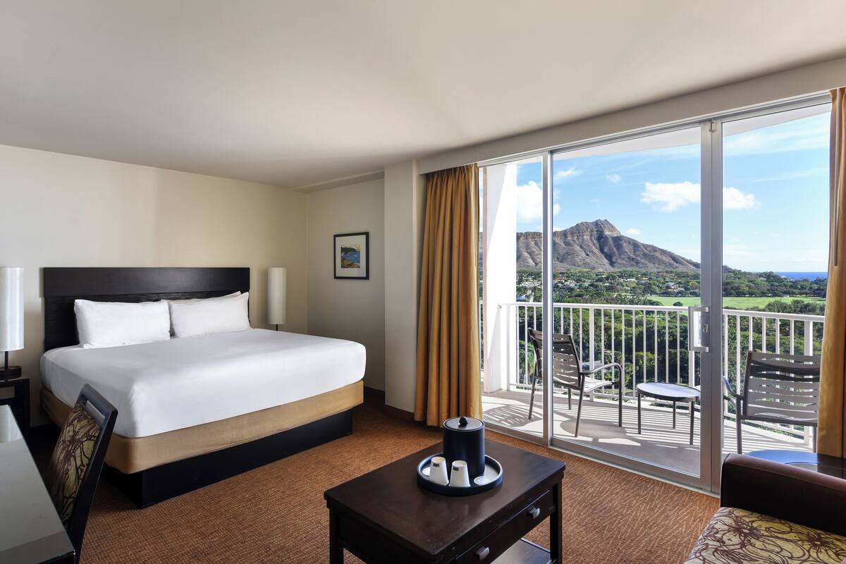 A hotel room with a large bed, a small table with a lamp, a chair, and a balcony with a view of a mountain and greenery.