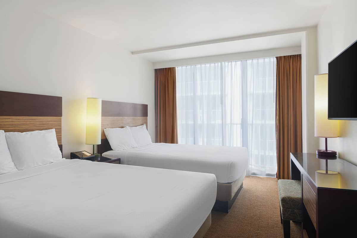 A hotel room with two beds, a desk, a TV, and a large window with curtains.