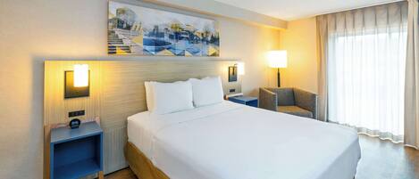 A hotel room with a large bed, two bedside tables, a chair, a painting on the wall, and a window with curtains.