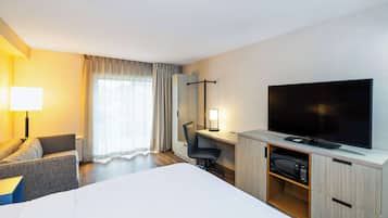 A hotel room with a bed, a desk with a flat-screen TV, a chair, a lamp, and a window with curtains.